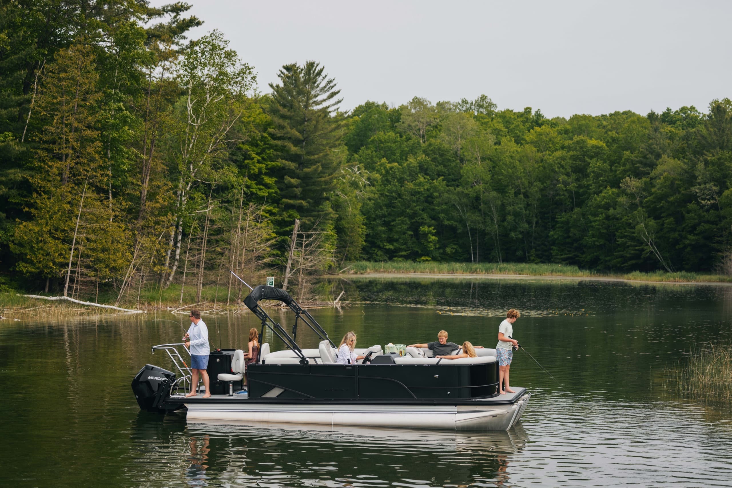Black-hull pontoon boat with guests fishing along a wooded shoreline